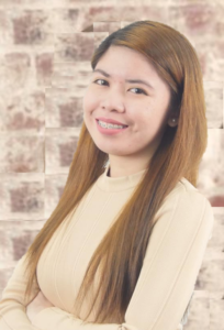 alt="Portrait of Iris Gueco, smiling against an exposed brick background"